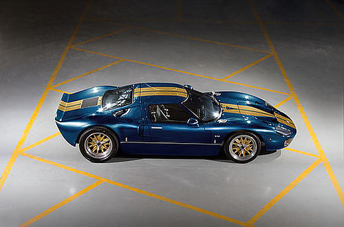 Low dark blue sports car with twin gold racing stripes and gold rims, shown from above on a concrete floor with yellow parking lines