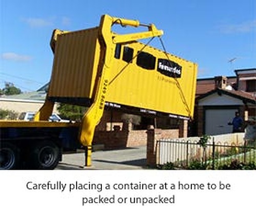 Yellow shipping container being lowered by a truck-mounted crane onto a suburban driveway beside a brick house.