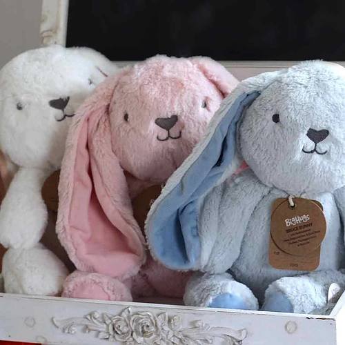 Three plush floppy-eared bunnies (white, pink, blue) in a carved white box, the blue one showing a brown tag.