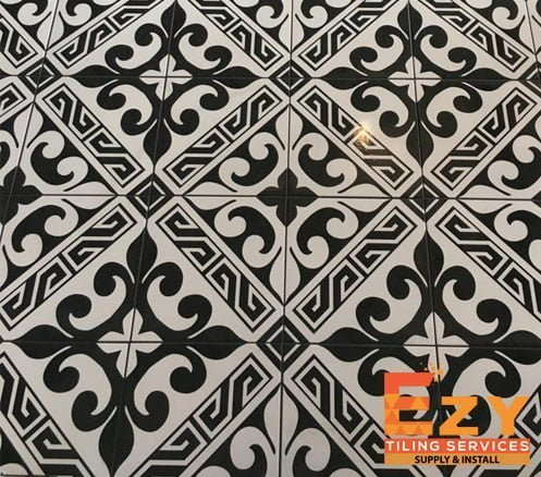 Black-and-white repeating geometric floor tiles with ornate scroll motifs and an orange Ezy Tiling Services logo in the lower-right corner