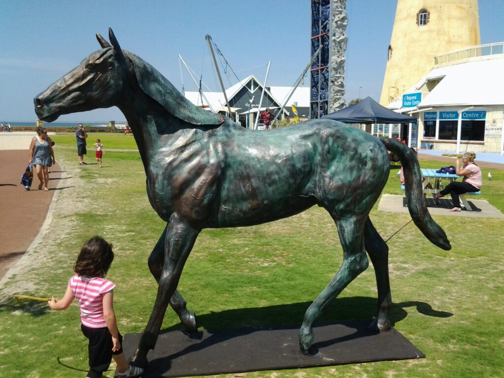 Life-size bronze horse sculpture with green patina in a walking pose on a grassy seaside promenade with people nearby