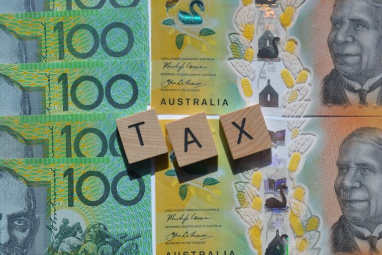 Australian $100 banknotes arranged beneath three wooden letter tiles spelling TAX.