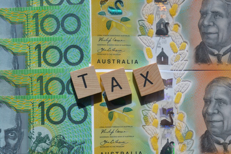 Australian $100 banknotes arranged beneath three wooden letter tiles spelling TAX.