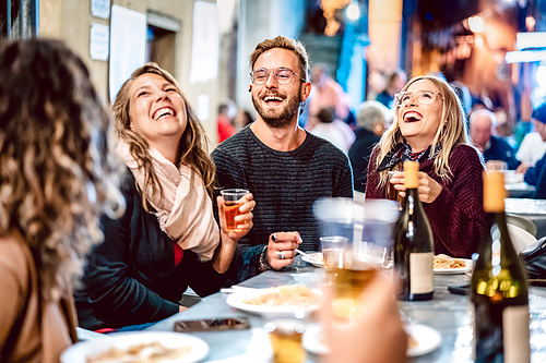 Group of friends laughing at an outdoor restaurant table at night, man with glasses holding a beer and wine bottles on table.