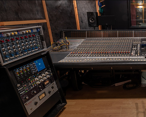 Large recording studio mixing console with many faders and knobs, adjacent rack of outboard processors and a monitor.
