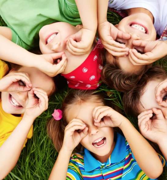 Five children lying on grass in a circle, smiling and making hand binoculars over their eyes.