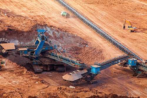 Open-pit mine with long blue conveyor belts feeding heavy earth-moving machinery and a yellow excavator on red-brown soil.