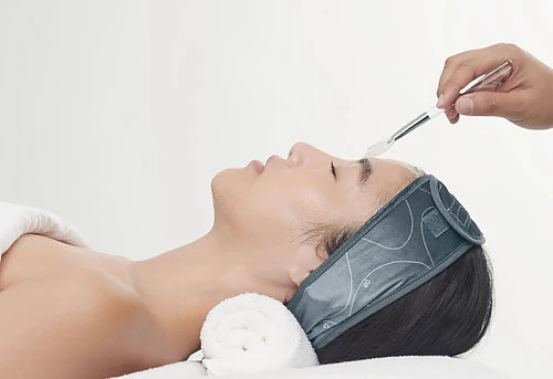 Woman wearing grey headband reclines on a rolled towel while a therapist brushes facial product onto her forehead