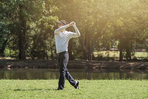 Golfer in white shirt and cap finishing a swing on grass by a pond with sunlit trees.
