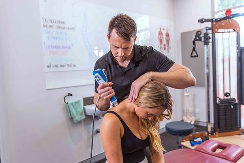 Physiotherapist using a handheld therapy device on a seated woman's neck in a clinic treatment room with exercise equipment.