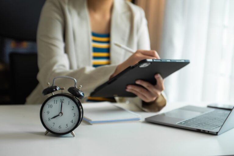 Black twin-bell alarm clock on a desk in the foreground with a blurred person using a tablet and laptop behind
