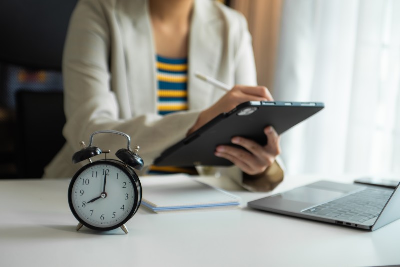 Black twin-bell alarm clock on a desk in the foreground with a blurred person using a tablet and laptop behind