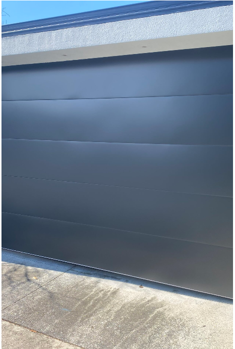 Smooth dark grey sectional garage door with horizontal panels beneath white stucco trim and a concrete driveway in front.