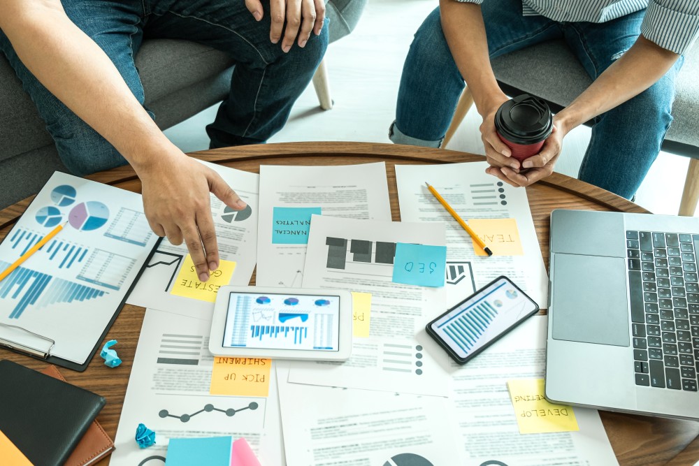 Two people at a round table reviewing charts on papers, tablet, phone and laptop with sticky notes and coffee.