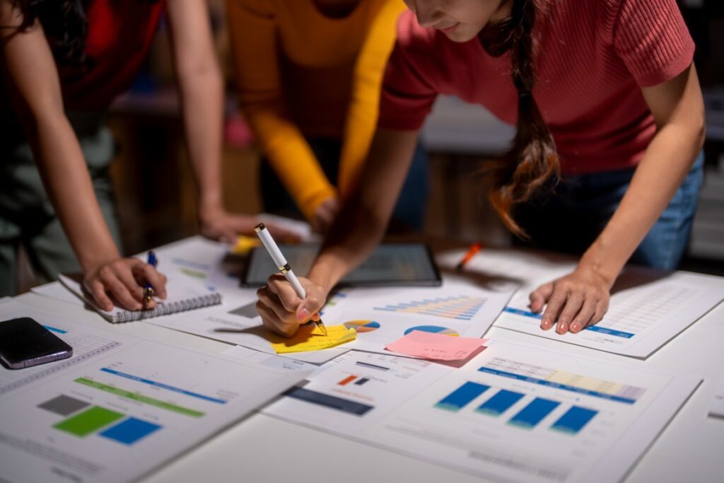 Group leaning over a table of charts and graphs, one person writing on a yellow sticky note while others consult a tablet.