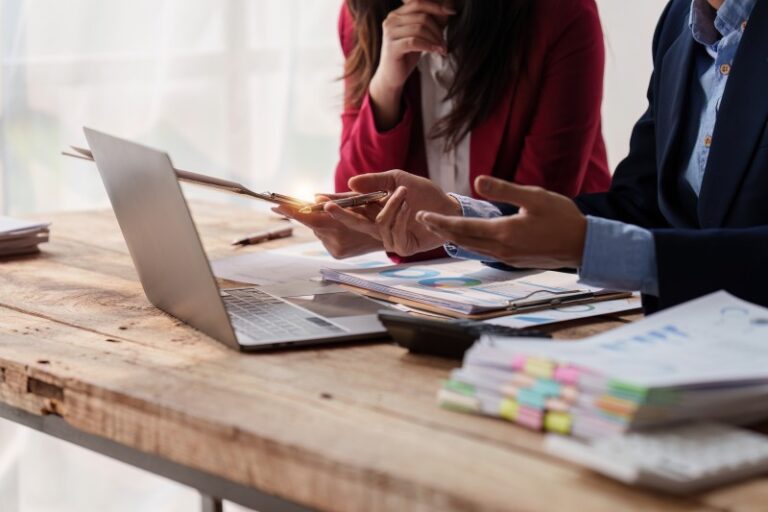 Two colleagues discussing charts and documents at a wooden table with an open laptop and stacked papers with coloured tabs.