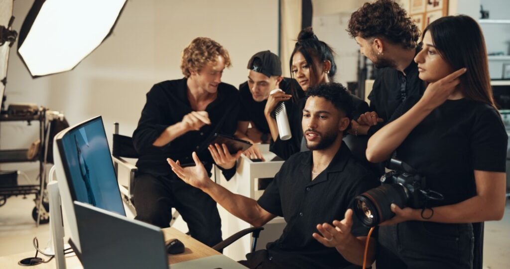 Studio crew gathered around a monitor reviewing photos, a seated man gesturing while a woman holds a camera and studio lights in the background.