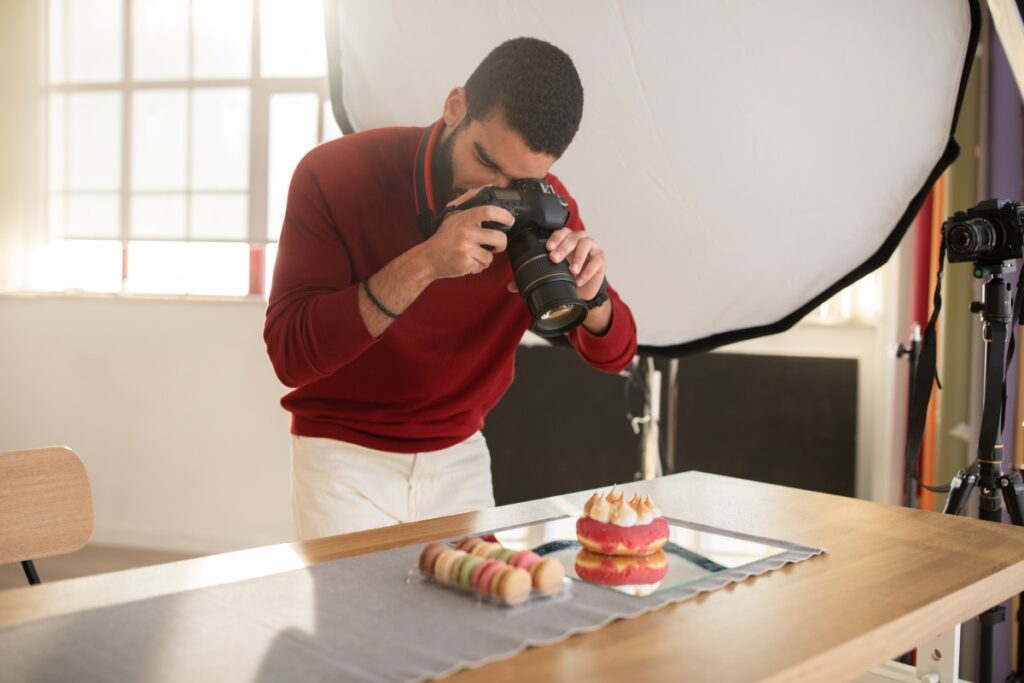 Photographer in red sweater using a DSLR to photograph pink cream-filled pastry and macarons on a table with studio softbox