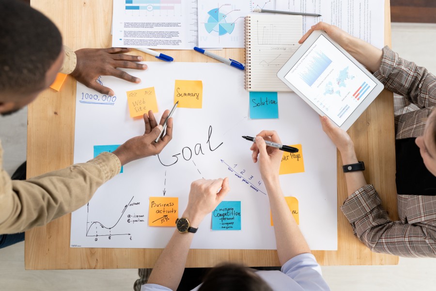 Overhead view of three people brainstorming around a table with Goal written on paper, sticky notes and tablet charts.