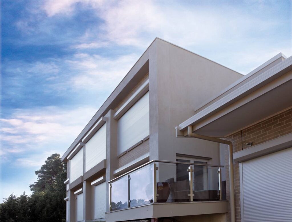 Modern two-storey house facade with glass balcony, white roller shutters and grey stucco under a blue sky.