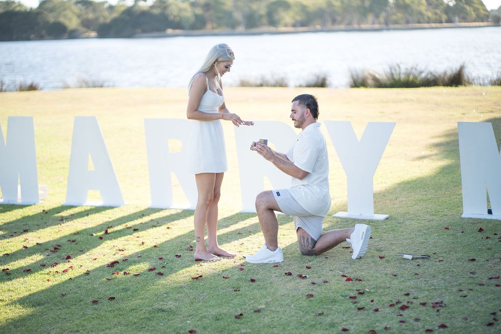 Man kneeling on grass proposing with ring to woman in white dress beside large white MARRY letters by a lakeside