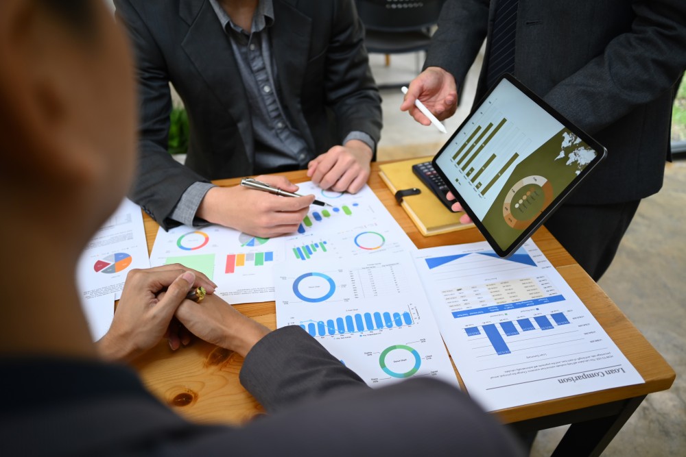 Three businesspeople in suits reviewing printed charts and a tablet displaying bar and pie graphs on a wooden table