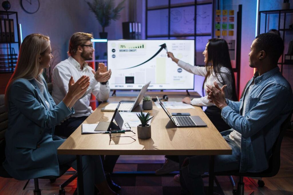 Four colleagues sit around a conference table, applauding as a woman points to an upward-trending chart displayed on a screen.