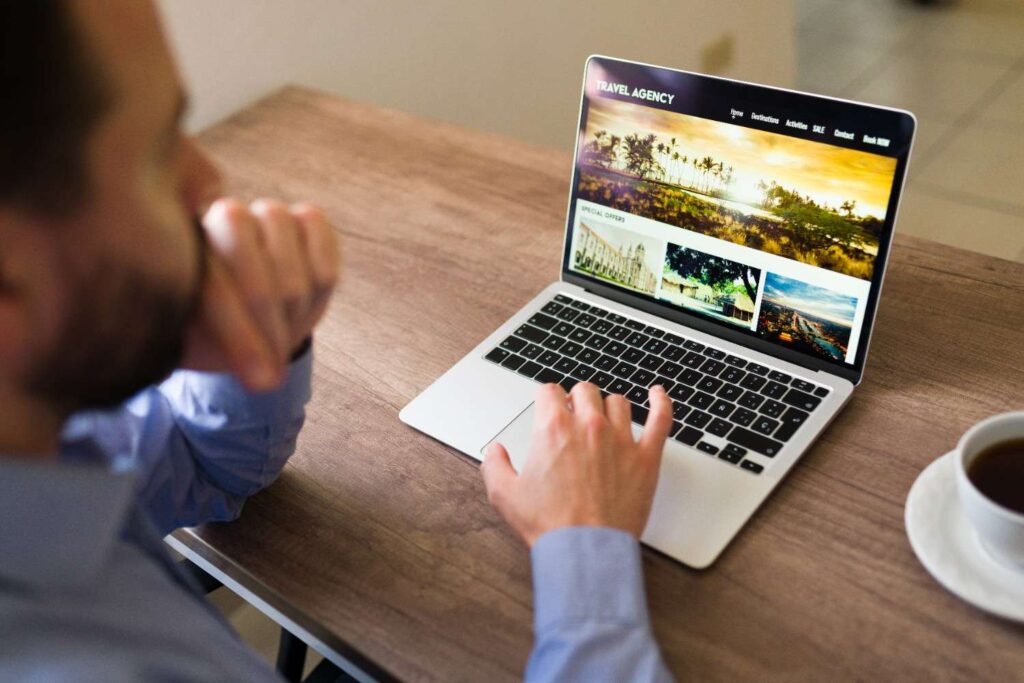 Person browsing a travel agency website on a laptop at a wooden table, with hand on the trackpad and a cup of coffee nearby.
