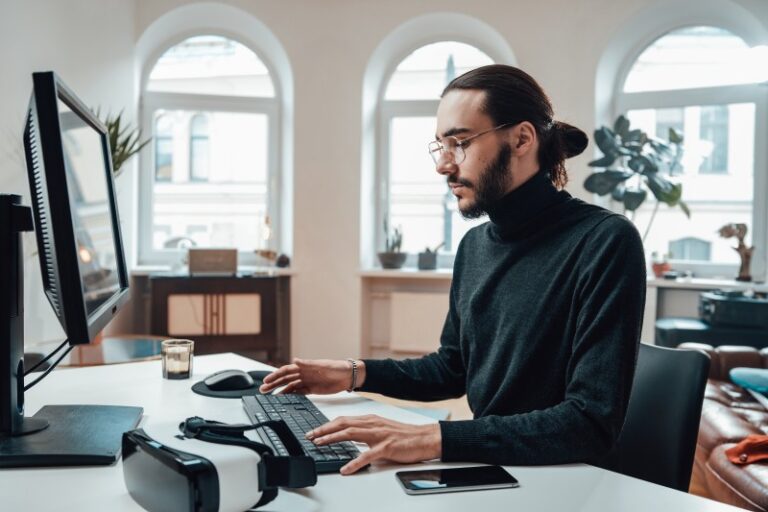 Man with glasses and hair in a bun typing at a keyboard in a bright office with arched windows; VR headset and phone on the desk.