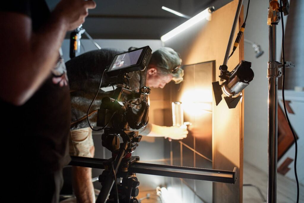 Filmmaker adjusting a glass prop against a lit panel while a camera on a tripod with monitor records the setup.