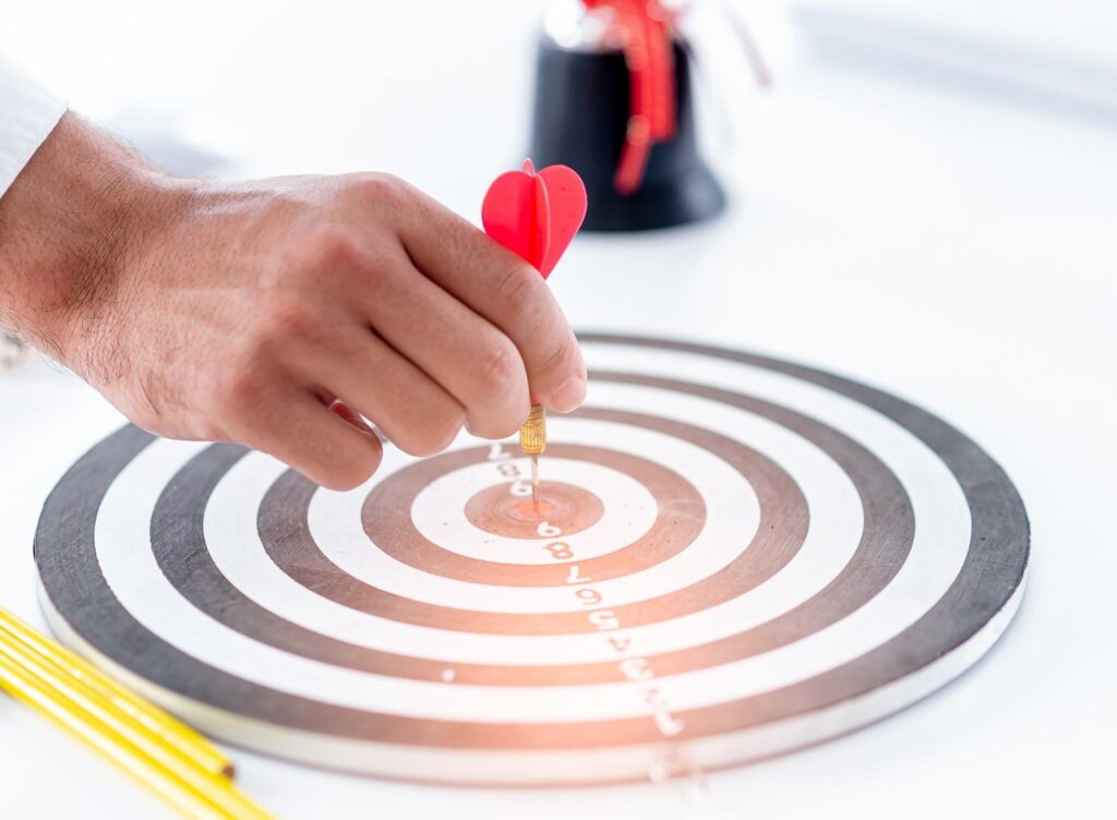 Hand placing a red heart-shaped dart into the bullseye of a black-and-white target on a white surface.