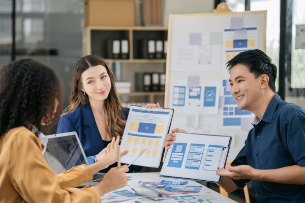 Three colleagues around a table sharing tablet wireframe mockups and printouts during a UX design meeting, smiling.