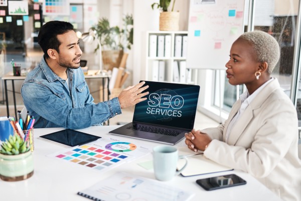 Colleagues in an office as a man shows a laptop screen reading SEO SERVICES, with colour swatches and devices on the table.