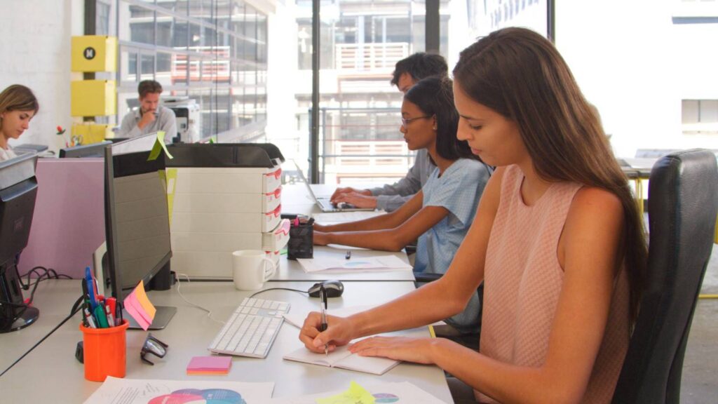 Young woman in sleeveless top writing in a notebook at an open-plan desk with colleagues using computers behind her.