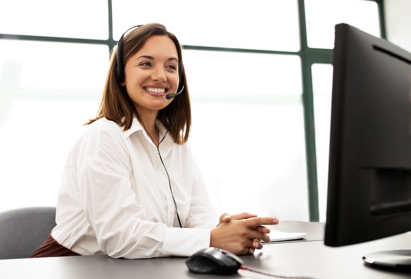 Smiling woman wearing a headset seated at a desk, hands clasped, looking toward a computer monitor in a bright office.