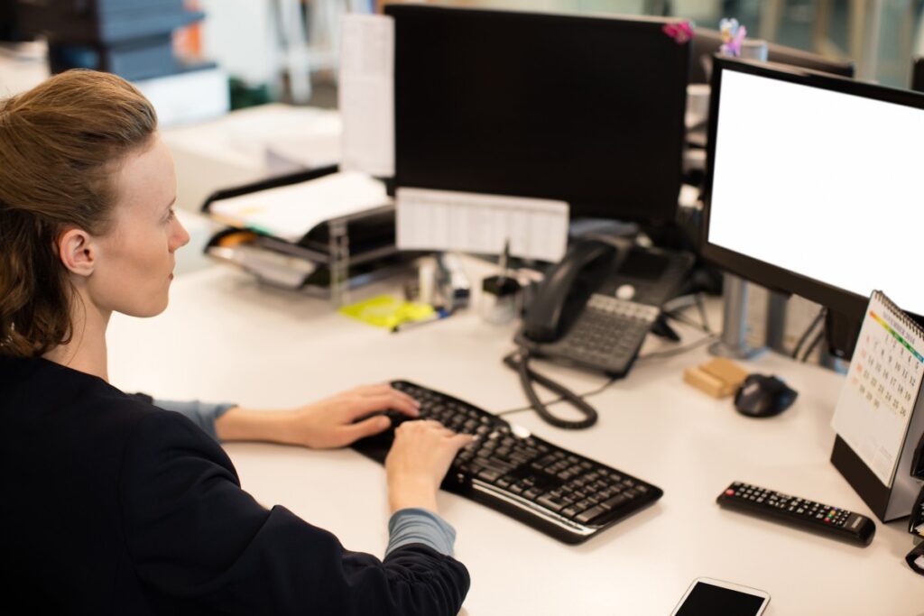 Woman typing at an office desk, using a keyboard with two computer monitors, telephone, calendar and smartphone nearby.