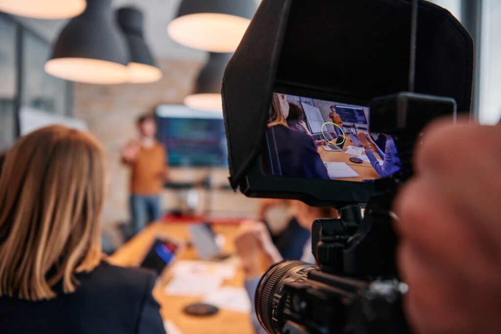 Camera filming a presenter and meeting participants around a conference table, shown on the camera's monitor.