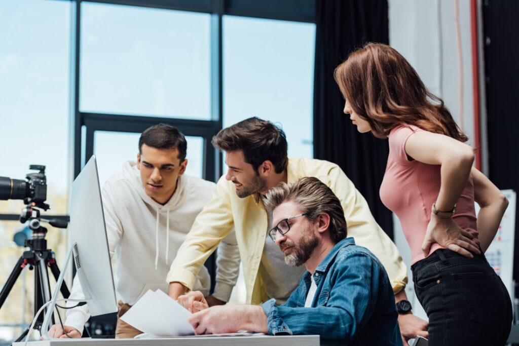 Four colleagues gathered around a desktop monitor in a bright studio, reviewing photos with a camera on a tripod.