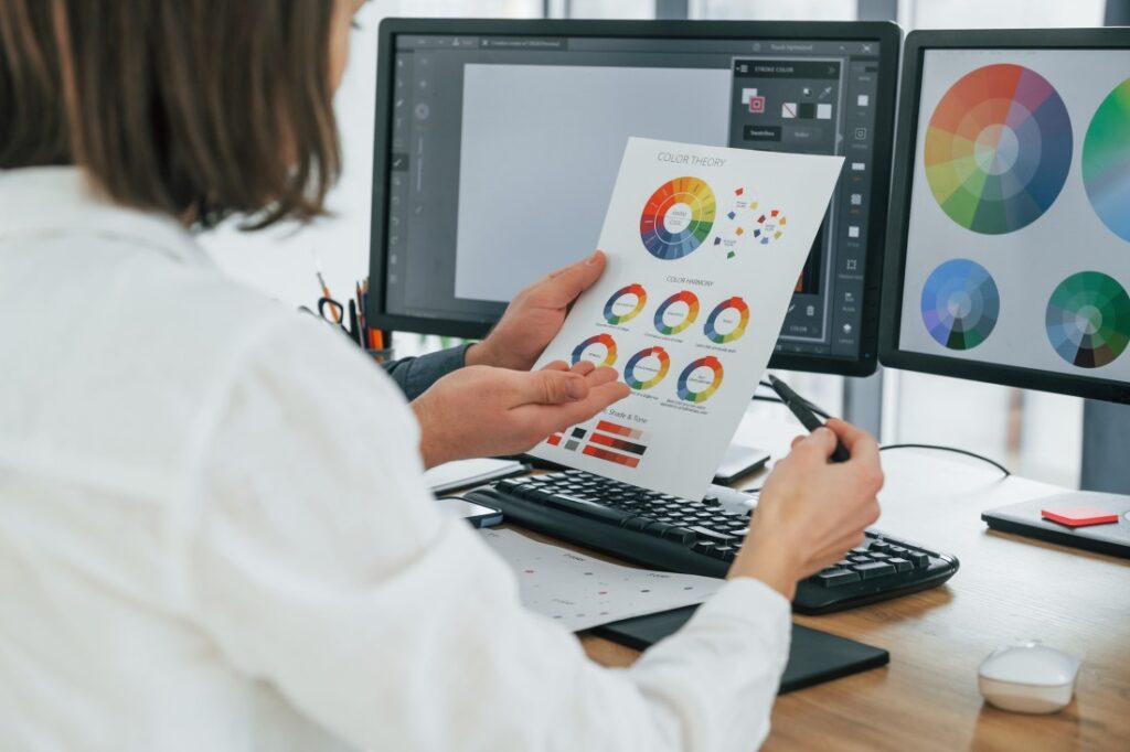 Graphic designer at a desk comparing a printed colour theory sheet with colour wheels to matching designs on dual monitors