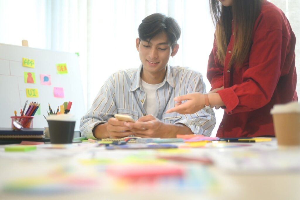 Young man smiling at his phone while a colleague in a red shirt points, amid sticky notes, sketches and stationery on a desk.