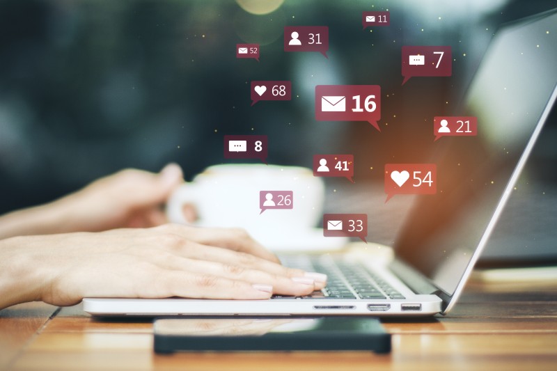 Hands typing on a laptop with floating social media notification icons (likes, messages, follower counts) above a coffee cup.