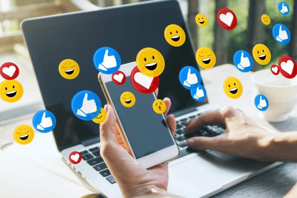 Hand holding smartphone over laptop with floating reaction emojis: blue thumbs, red hearts and yellow smileys.