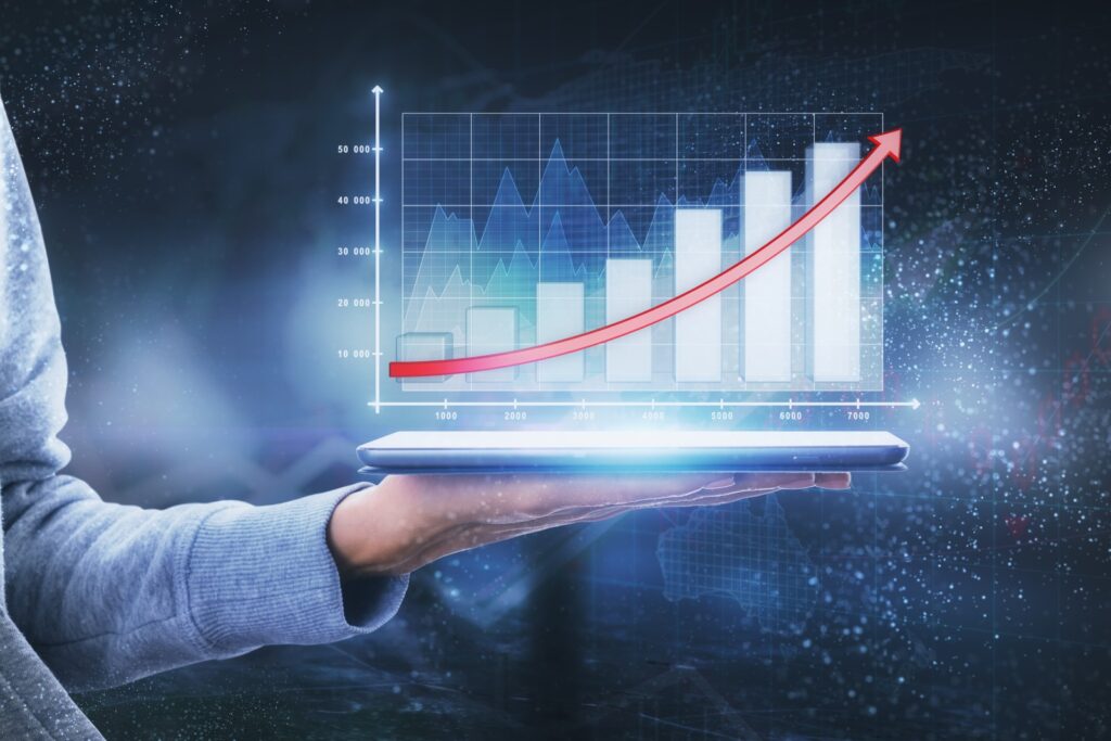 Hand holding a tablet projecting a bar chart with rising bars and a red upward arrow on a blue background.