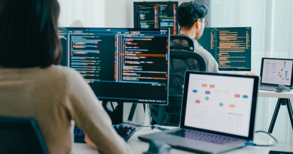 Two software developers at desks working on multiple monitors displaying source code and a laptop showing app diagrams.