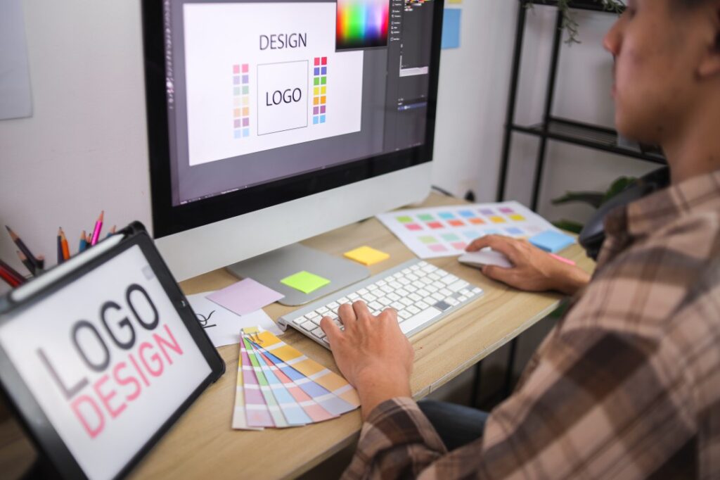 Designer at a desk creating a logo on a computer with colour swatches and a tablet showing the words LOGO DESIGN.