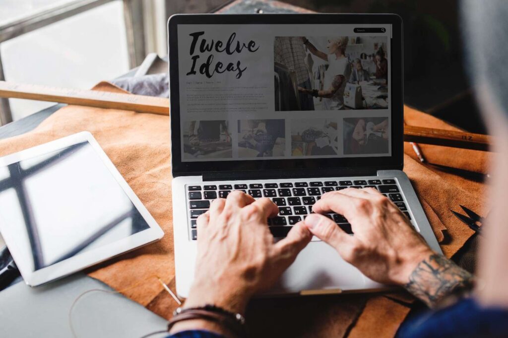 Tattooed hands typing on a laptop displaying a "Twelve Ideas" webpage, with a tablet and tools on a leather workspace.