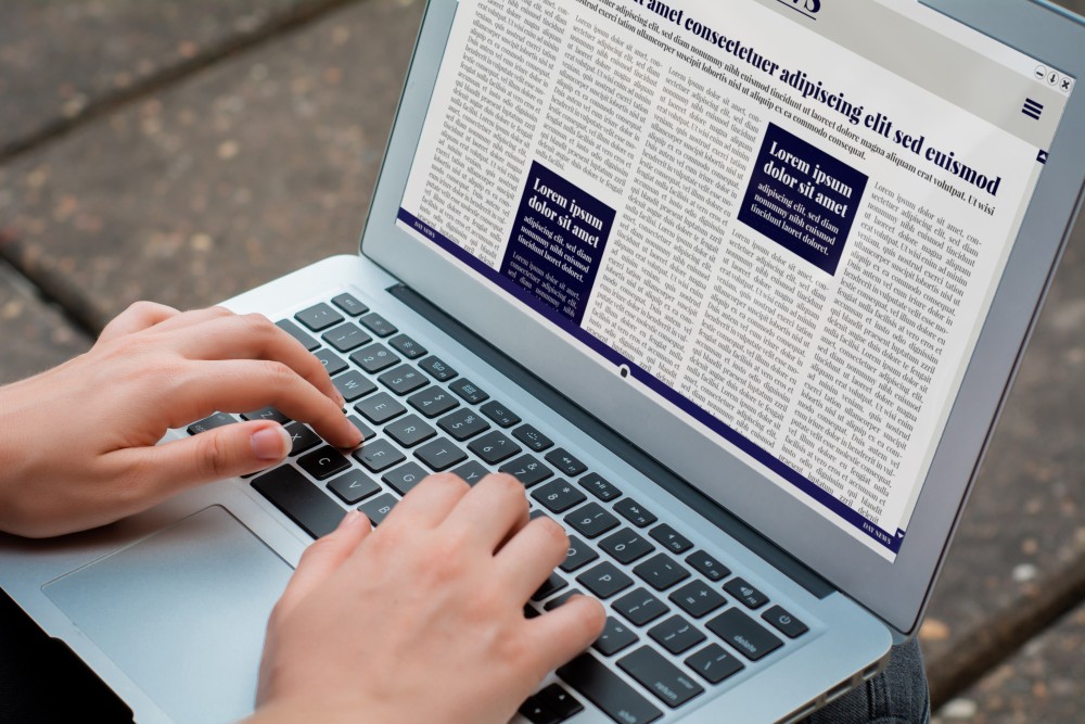 Hands typing on a silver laptop keyboard displaying a multi-column digital newspaper page with blue callout boxes.