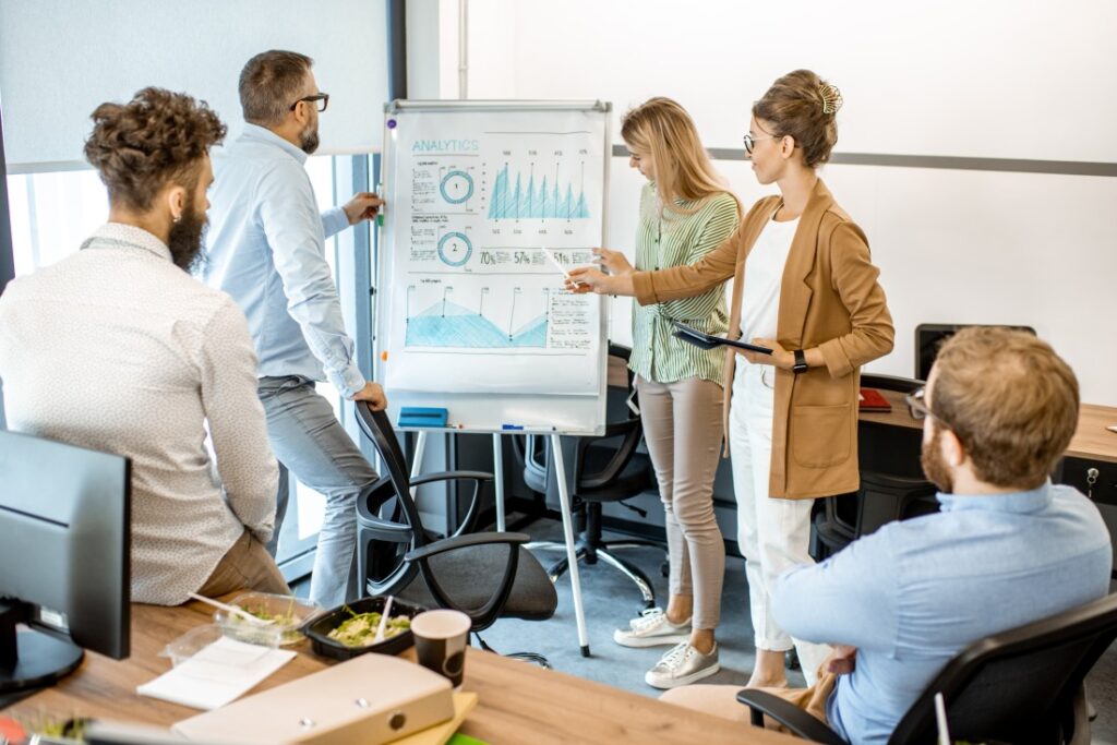 Five colleagues in a modern office gathered around a flip chart with blue analytics graphs as one presents.