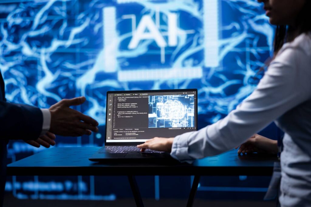 Laptop showing code and a glowing digital brain graphic, with two people gesturing beside it in a blue AI-themed room