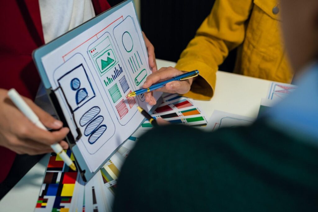 People pointing with pens at a clipboard of colourful mobile app wireframe sketches beside printed colour swatches.